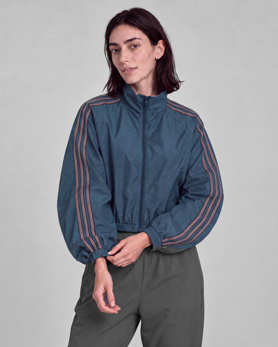Woman in a teal blue cropped track jacket with three taupe sleeve stripes and gray trousers.