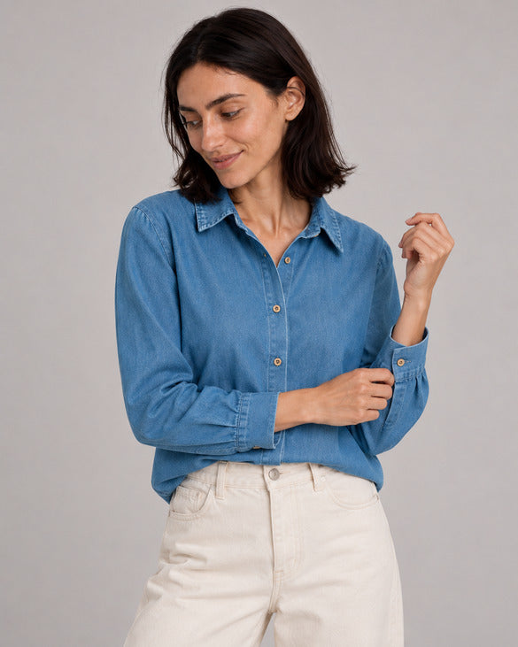 Blue relaxed denim shirt on a model adjusting one cuff, paired with ivory jeans.