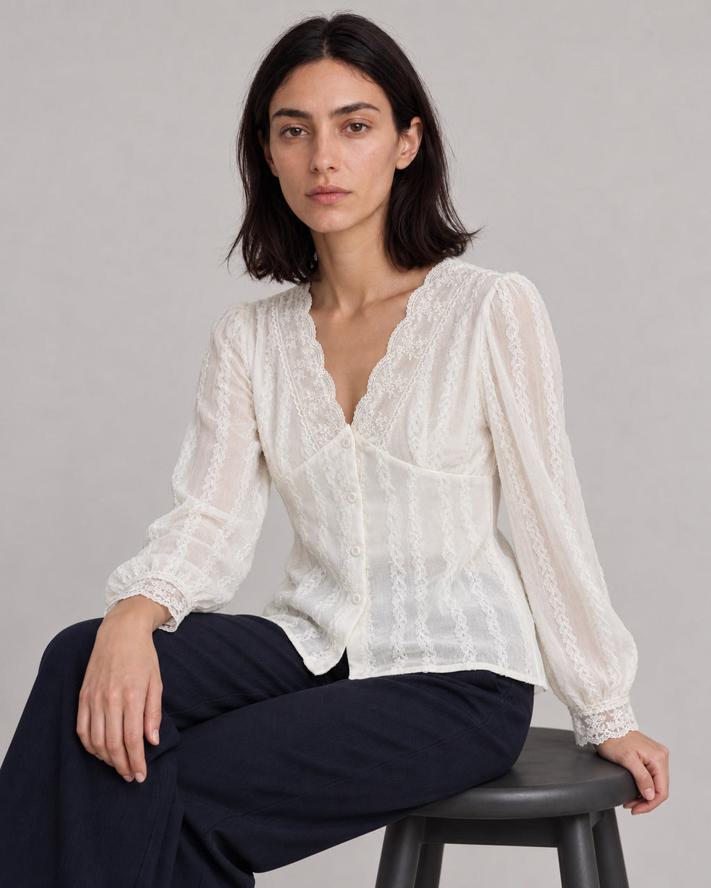 White lace-trim V-neck blouse on a model seated sideways on a gray stool.